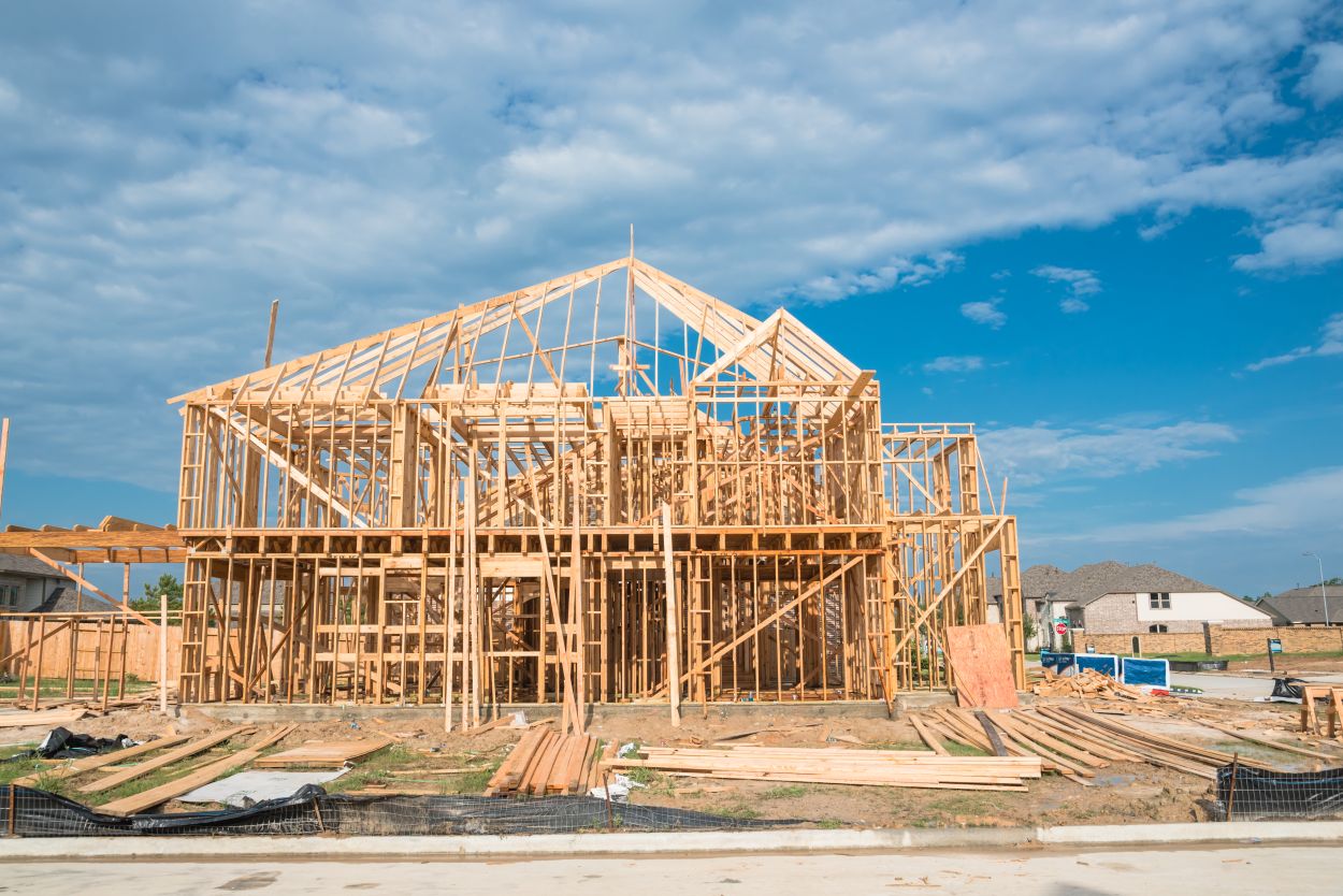 New stick built home under construction and blue sky in US. Framing structure/wood frame of wooden houses next to completed suburban home. Pile of sand, gravel, logs in front. Industrial, real estate See Less