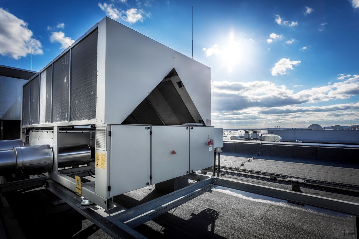 A huge air conditioning unit on the roof of the building. In the background of blue sky with shining sun. Focus is at the front of the air conditioner, the other parts of image slightly ... See More